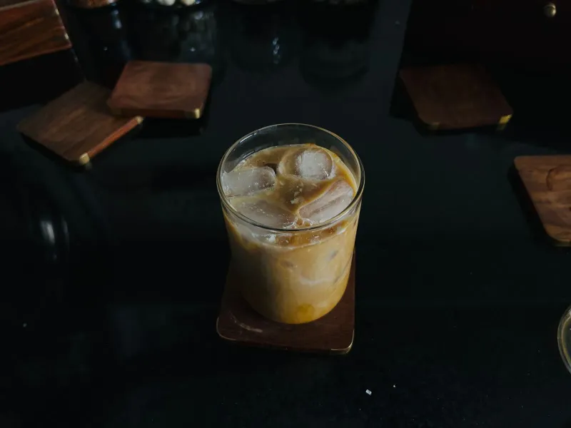 Un verre de boisson glacée ambrée sur un sous-verre en bois, avec quelques cosses séchées éparpillées sur la table sombre