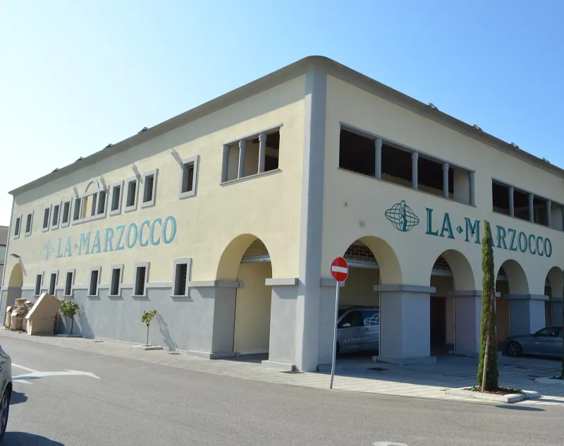 Cream-coloured La Marzocco factory building with arches and the La Marzocco name on its facade, blue sky above and cars parked in front