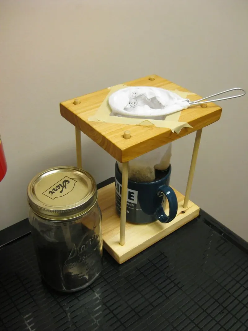 Weißer Kaffeebeutel aus Stoff auf einem Holzgestell über einem blauen Becher, daneben ein Glas mit gemahlenem Kaffee