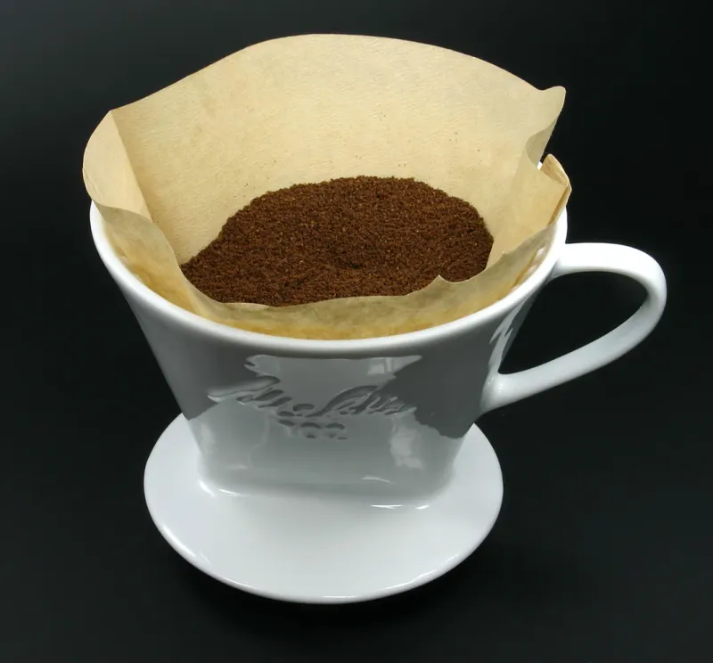 Weißer Melitta-102-Filterhalter aus Porzellan mit einer braunen Papierfiltertüte voller gemahlenem Kaffee