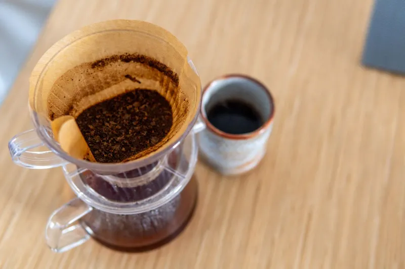 Gerippter, durchsichtiger Kegel-Dripper mit gefaltetem Papierfilter voller Kaffeesatz neben einem kleinen Becher auf einem Holztisch
