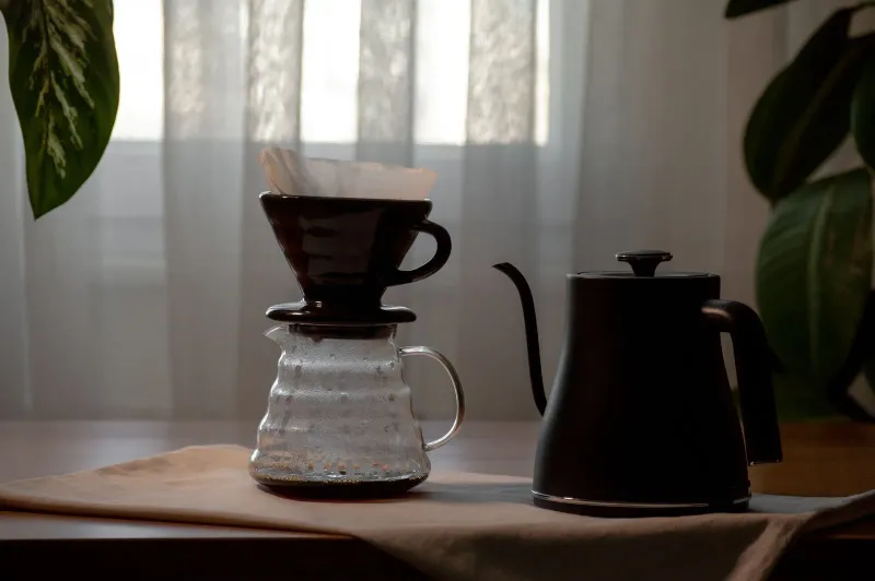 Schwarzer Keramik-Kegel-Dripper mit leerem weißem Papierfilter auf einer Glaskaraffe, daneben ein schwarzer Schwanenhals-Wasserkocher