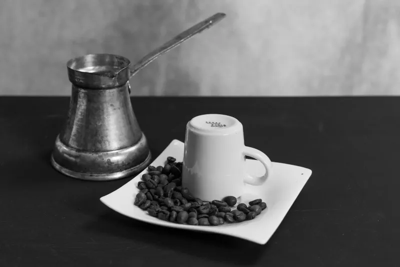 Schwarz-Weiß-Stillleben eines Cezve mit langem Stiel neben einer kleinen Tasse mit Untertasse, umgeben von Kaffeebohnen