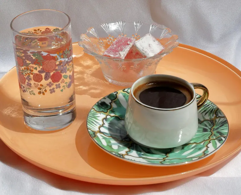 Kleine grüne Porzellantasse mit schaumigem türkischem Kaffee auf einem orangefarbenen Tablett, mit einem Glas Wasser und rosa und weißem Lokum
